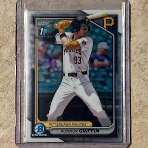 Puede incluir: Tarjeta de béisbol de Konnor Griffin de los Piratas de Pittsburgh, en una funda protectora. La tarjeta tiene un acabado cromado y presenta el logo del equipo, el nombre del jugador y "1st Bowman". El jugador viste un uniforme blanco y negro con el número 93.