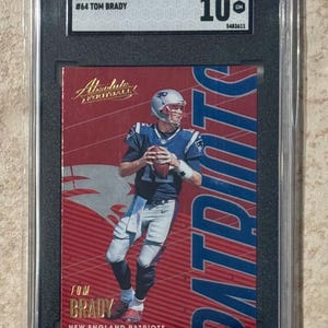 Puede incluir: Una tarjeta de fútbol Panini Absolute 2018 calificada con Tom Brady de los New England Patriots. La tarjeta tiene un fondo rojo con la palabra "PATRIOTS" en azul. La tarjeta está calificada con un 10.