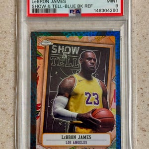May include: A 2024 Topps Chrome LeBron James Show & Tell Blue BK Refractor trading card graded PSA Mint 9. The card features LeBron James in a yellow Los Angeles jersey with number 23, holding a basketball in front of a chalkboard background.