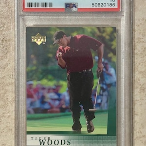 Puede incluir: Tarjeta coleccionable Upper Deck Golf 2001 con Tiger Woods. La tarjeta, calificada GEM MT 10, está en un soporte protector. La imagen muestra a Woods en un campo de golf, con camisa granate y pantalón negro.