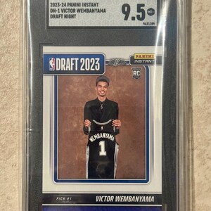 May include: A 2023-24 Panini Instant Victor Wembanyama Draft Night trading card graded 9.5 by SGC. The card features a photograph of Victor Wembanyama holding a black San Antonio Spurs jersey with the name Wembanyama and number 1 on the back.