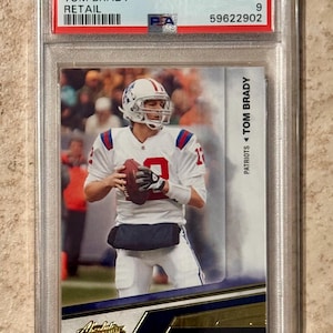 Puede incluir: Una tarjeta de fútbol Tom Brady 2010 Panini Absolute Memorabilia, calificada por PSA. La tarjeta presenta a Brady con su uniforme de los Patriots, sosteniendo un balón de fútbol. La tarjeta está dentro de un soporte de plástico transparente.