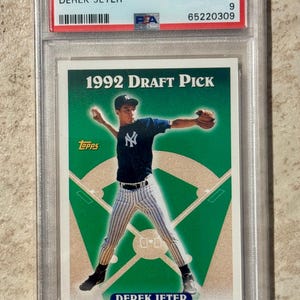 May include: A 1993 Topps Derek Jeter baseball card, graded Mint 9 by PSA, encased in a protective holder. The card features a 1992 Draft Pick image of Jeter in a Yankees uniform, throwing a baseball on a green background.