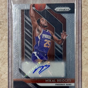 May include: A basketball trading card featuring Mikal Bridges of the Phoenix Suns in a purple and orange jersey, mid-jump with a basketball. The card is encased in a protective sleeve, with the "PRIZM" logo visible.