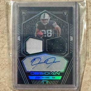 May include: A football trading card featuring Josh Jacobs of the Raiders. The card has a black and silver design with a holographic signature and the word "OBSIDIAN". The card is encased in a protective sleeve.