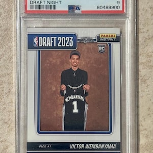 May include: A 2023 Panini Instant Victor Wembanyama Draft Night trading card graded Mint 9 by PSA. The card features a photograph of Victor Wembanyama holding a black San Antonio Spurs jersey with the number 1, encased in a clear plastic slab.