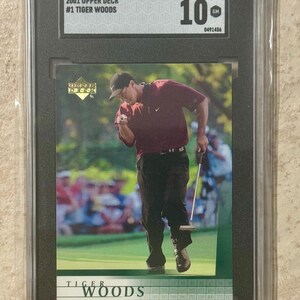 Puede incluir: Una tarjeta comercial de Upper Deck de 2001 calificada con Tiger Woods. La tarjeta está dentro de un soporte negro y tiene una calificación de 10. La imagen muestra a Tiger Woods en un campo de golf, celebrando un golpe.