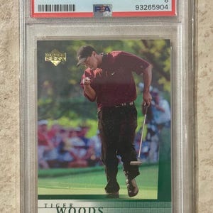 May include: A 2001 Upper Deck Golf trading card featuring Tiger Woods. The card is graded NM-MT 8 by PSA and shows Woods celebrating on the course. The card is encased in a protective holder.