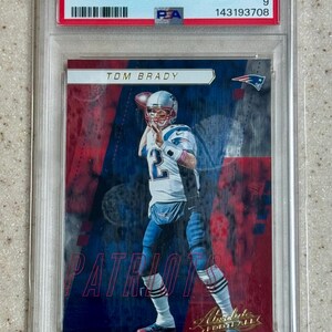 Puede incluir: Una tarjeta de fútbol americano 2017 Panini Absolute de Tom Brady, clasificada como PSA Mint 9. La tarjeta muestra a Tom Brady con uniforme blanco y azul lanzando un balón, sobre un fondo rojo y azul con el texto "Tom Brady" y "Patriots".
