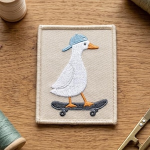 May include: A rectangular fabric patch featuring an embroidered white duck wearing a light blue baseball cap and standing on a dark gray skateboard. The duck has an orange beak and orange feet, set against a light beige background.