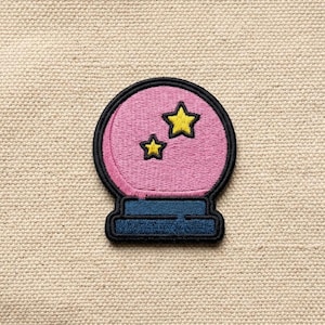 May include: Embroidered patch featuring a pink crystal ball with two yellow stars. The crystal ball has a black outline and sits on a blue base. The patch is designed for clothing or accessories.