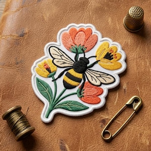 May include: An embroidered patch featuring a central black and yellow bee surrounded by orange and yellow flowers with green leaves. The patch is accompanied by a small metal thimble, a spool of olive green thread, and a gold-toned safety pin.