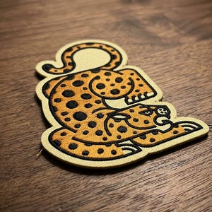 May include: Embroidered patch featuring a stylized leopard in shades of orange and black. The leopard is depicted in a crouching pose with its tail curled. The patch has a tan border and is likely for clothing or accessories.