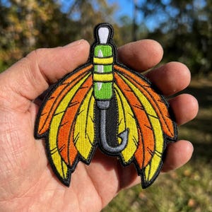 May include: An embroidered patch featuring a central green and yellow fishing float with a white top, attached to a grey fishing hook. The design is flanked by symmetrical, layered feathers in vibrant shades of orange and yellow with black outlines.