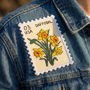 May include: A rectangular embroidered patch shaped like a postage stamp featuring yellow daffodils with orange centers on a cream background. The text 03 USA DAFFODIL is stitched in black thread. The patch has a white scalloped border.