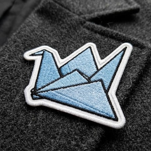 May include: A light blue origami crane patch with a white border and black outline is sewn onto a dark gray fabric. The patch is a geometric design of a folded paper crane.