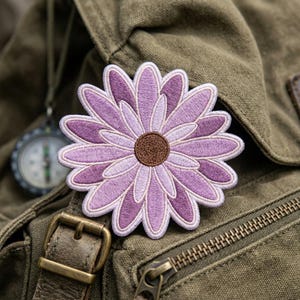 May include: Embroidered daisy patch in shades of purple and white, with a brown center. The flower is affixed to a green canvas bag with a brass buckle and zipper. A compass hangs in the background.