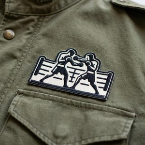 May include: Olive green jacket with a boxing patch. The patch features two boxers in a ring, rendered in black and white. The jacket has a button closure and a pocket on the front.