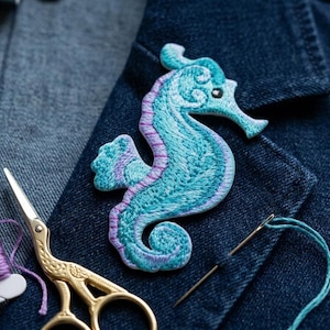 May include: An embroidered seahorse patch in shades of teal and purple rests on a denim jacket. Gold scissors, a spool of purple thread, and a needle with teal thread are also visible, suggesting a crafting theme.