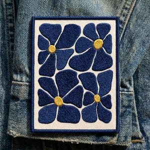 May include: A rectangular patch featuring a floral design. The patch has a white border and background. The flowers are a deep blue with yellow centers. The patch is attached to a denim jacket.