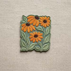 May include: Embroidered patch featuring orange coneflowers with brown centers and green leaves on a teal background. The patch has a green border and is set against a neutral linen backdrop. A floral design.