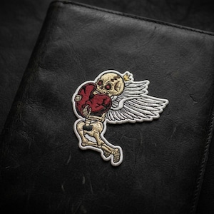 May include: Embroidered patch featuring a skeleton angel embracing a red heart. The patch has a cream-colored skeleton with a gold crown, white wings, and a red heart. The patch is on a black leather surface.
