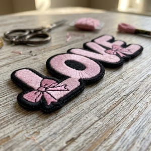 May include: A pink embroidered patch with the word "LOVE" in a bubble letter style, featuring bow accents. The patch has a black border and is set against a light wooden surface. Sewing supplies, including scissors and thread, are in the background.
