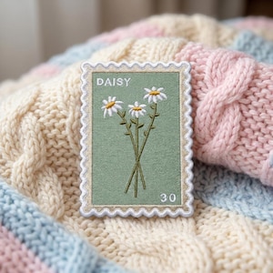 May include: A rectangular embroidered patch shaped like a postage stamp with a scalloped edge. The patch features a sage green background with three white daisy flowers with yellow centers, the text DAISY, and the number 30 in white.