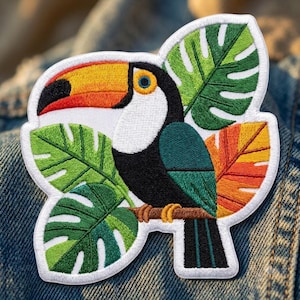 May include: An embroidered patch featuring a toucan with a large orange, yellow, and black beak, a white chest, and black and teal feathers. The bird is perched on a brown branch surrounded by green monstera leaves and a single orange leaf.
