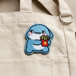 May include: An embroidered patch featuring a light blue shark character with a white belly and pink cheeks. The shark is holding a red container filled with yellow french fries. The patch is attached to a beige canvas tote bag with a metal ring.