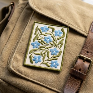 May include: Embroidered patch featuring blue flowers and green leaves on a cream background, framed by a green border. The patch is affixed to a tan canvas bag with a brown leather strap and buckle.