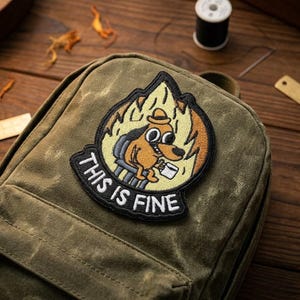 May include: Olive green backpack with a humorous embroidered patch. The patch features a cartoon dog in a burning room, holding a coffee cup, with the text "THIS IS FINE" in white letters.
