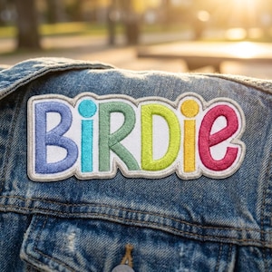 May include: A rectangular embroidered patch featuring the word BIRDIE in colorful, individual letters. The B is light blue, the I is turquoise, the R is sage green, the D is lime green, the I is yellow, and the E is magenta, all on denim.