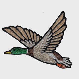 May include: Embroidered patch depicting a flying duck with open wings. The duck is coloured in shades of brown, tan, green, and orange, with black edging. The patch is designed for sewing or ironing onto fabric.