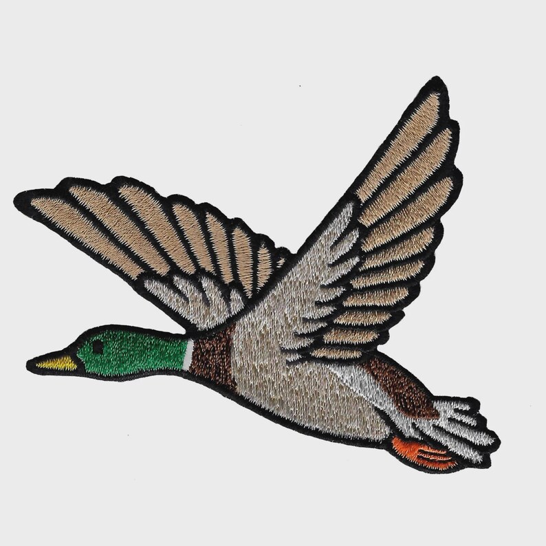 May include: Embroidered patch depicting a flying duck with open wings. The duck is coloured in shades of brown, tan, green, and orange, with black edging. The patch is designed for sewing or ironing onto fabric.