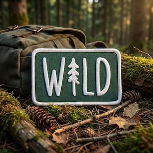 May include: A rectangular embroidered patch with a dark green background and a white border. The patch features the white text WILD, where the letter I is replaced by a stylized white pine tree graphic, centered on the fabric surface.