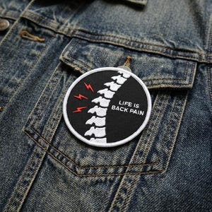 May include: A circular embroidered patch with a black background, featuring a white human spine illustration, three red lightning bolt symbols, and the white text "LIFE IS BACK PAIN" stitched onto a blue denim fabric surface.