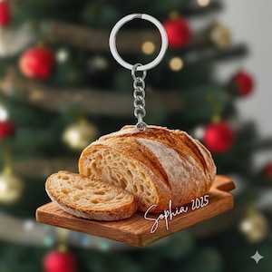 May include: A bread-shaped keychain featuring a loaf of crusty bread and a slice on a wooden cutting board. The keychain has a silver ring and chain. The name "Sophia 2025" is written on the board.