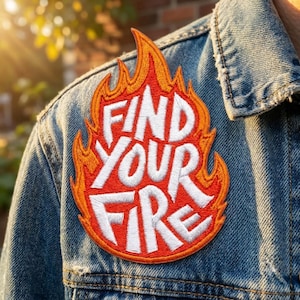 May include: An embroidered patch attached to a blue denim jacket. The patch is shaped like a flame with orange and red stitching, featuring the white text "FIND YOUR FIRE" in a bold, stylized font centered within the flame design.