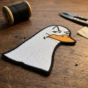 May include: Embroidered patch featuring a grumpy goose head with an orange beak and black outline. A spool of black thread and scissors are also visible. The patch is white with a black border.
