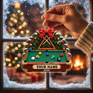 May include: A flat, decorative Christmas ornament shaped like a pool table with a green felt surface, billiard balls, and a wooden frame. Above the table is a pine wreath with a red bow and crossed pool cues. The text reads YOUR NAME and 2026.