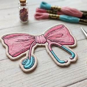 May include: Embroidered patch featuring a pink bow and stethoscope design. The patch has a cream-colored border and is accented with light blue details. A small glass bottle with dried flowers, embroidery floss, and gold scissors are in the background.