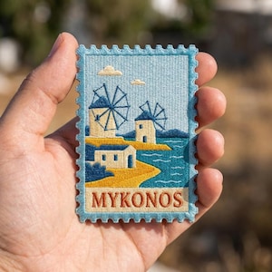 May include: A rectangular embroidered patch shaped like a postage stamp featuring a scene of Mykonos with two windmills, white buildings, a yellow coastline, and blue water under a light blue sky. The word MYKONOS is stitched in orange at the bottom.