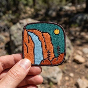 May include: A square embroidered patch with rounded corners featuring a landscape scene. The design depicts a light blue waterfall cascading down orange cliffs against a dark teal sky with a yellow sun and two small black pine tree silhouettes.