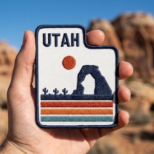 May include: A rectangular embroidered patch with a white background and a dark blue border. The text UTAH is at the top in dark blue. Below is a dark blue silhouette of a natural rock arch, three small cacti, an orange sun, and three horizontal stripes in red, orange, and teal.