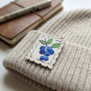 May include: A beige knit beanie with a decorative patch featuring embroidered blueberries and green leaves. The patch is attached with a safety pin. A brown leather notebook and pen are in the background.