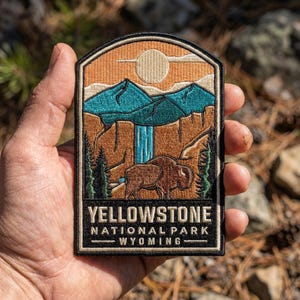 May include: An embroidered patch featuring a landscape of Yellowstone National Park in Wyoming. The design includes a brown bison, a blue waterfall, teal mountains, and a tan sun against an orange sky. The text reads YELLOWSTONE NATIONAL PARK WYOMING.