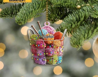 May include: A 2D flat ornament shaped like a wire basket filled with colorful yarn balls and knitting needles. The basket is adorned with string lights and the name "Sophia". The ornament hangs from a gold chain, perfect for a Christmas tree.