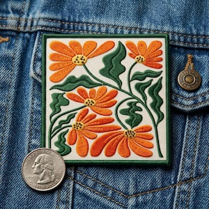 May include: Embroidered square patch featuring orange flowers with yellow centers and green leaves on a white background. The patch has a green border and is displayed on a denim jacket. A quarter coin is next to the patch.