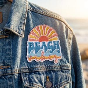 May include: An embroidered patch attached to a blue denim jacket. The patch features a stylized sun in shades of coral and yellow above the blue text "BEACH" and the word "vibes" in cursive, set against a background of light blue ocean waves.